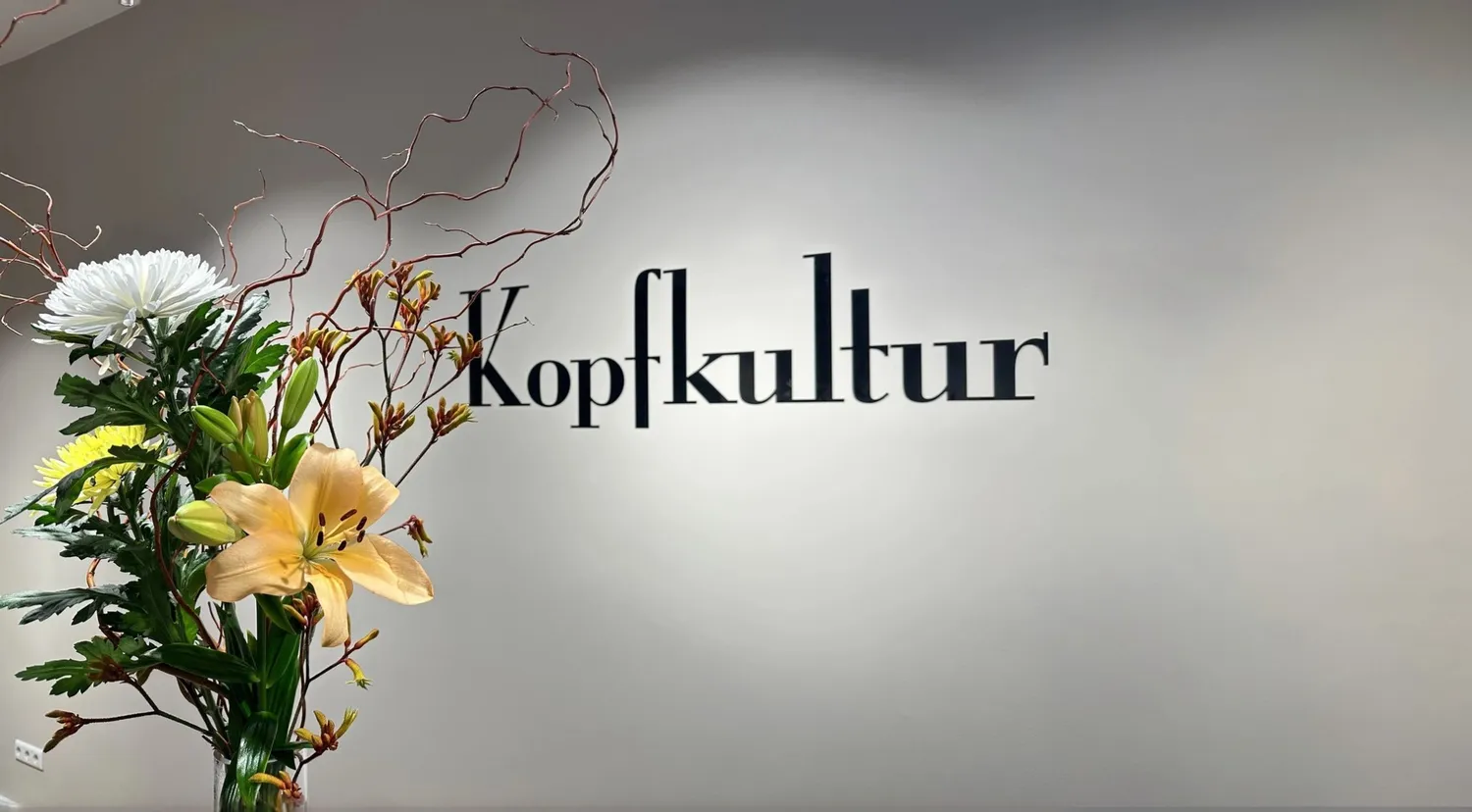 Lukas Ramolla – Haarstudio – Auf dem Bild ist das Wort „Kopfkultur“ in stilisierter Serifenschrift vor einer neutralen Wand zu sehen, begleitet von einem dekorativen Blumenarrangement.