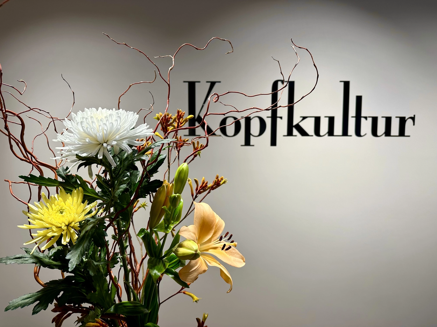 Lukas Ramolla – Haarstudio – Auf dem Bild ist das Wort „Kopfkultur“ in stilisierter Serifenschrift vor einer neutralen Wand zu sehen, begleitet von einem dekorativen Blumenarrangement.